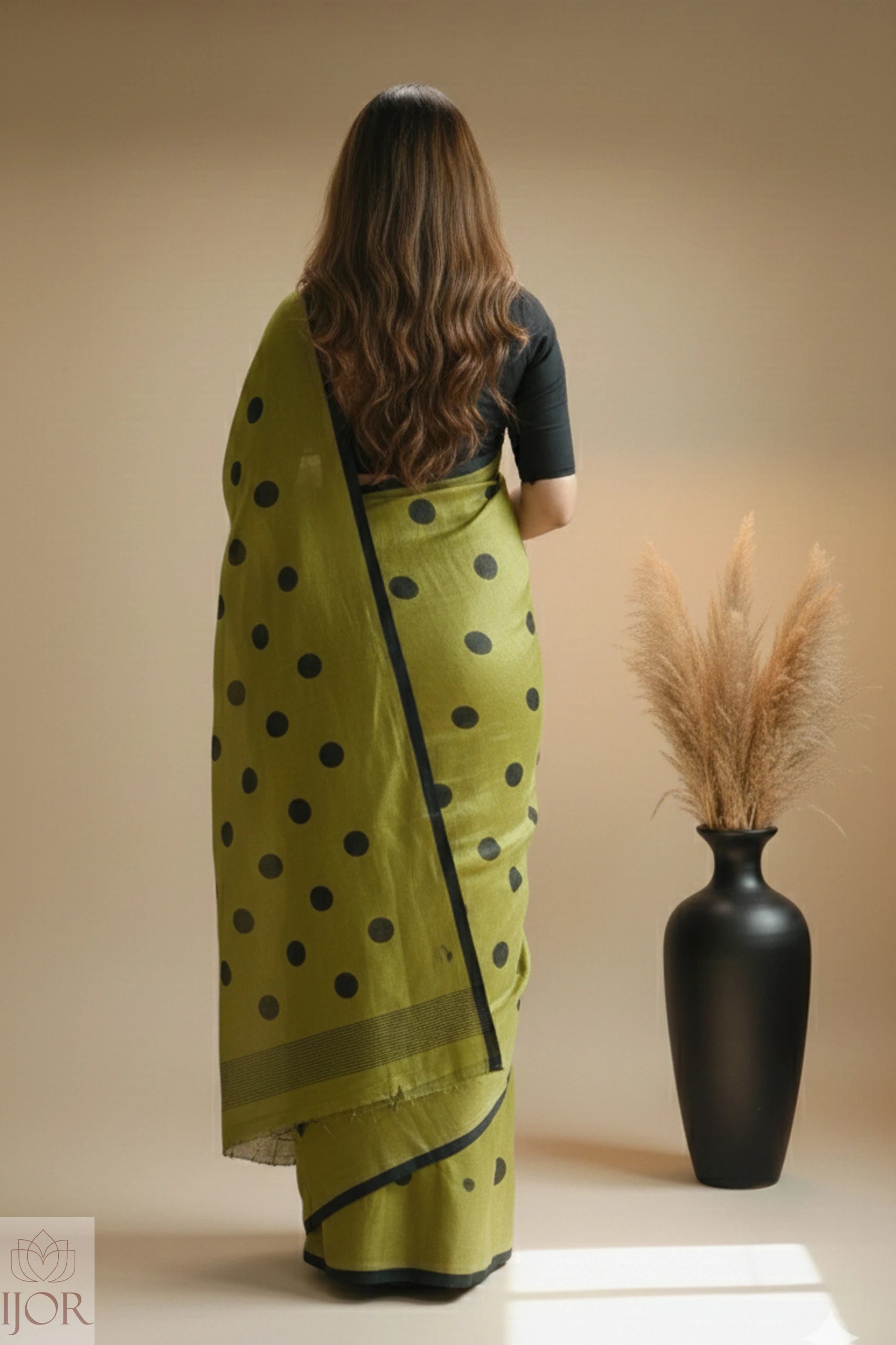 Pure Khadi Cotton Olive Green Saree with black polka, standing in front of a black vase with dried plants on a beige background.