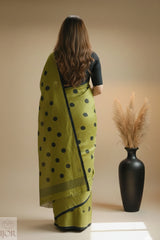 Pure Khadi Cotton Olive Green Saree with black polka, standing in front of a black vase with dried plants on a beige background.
