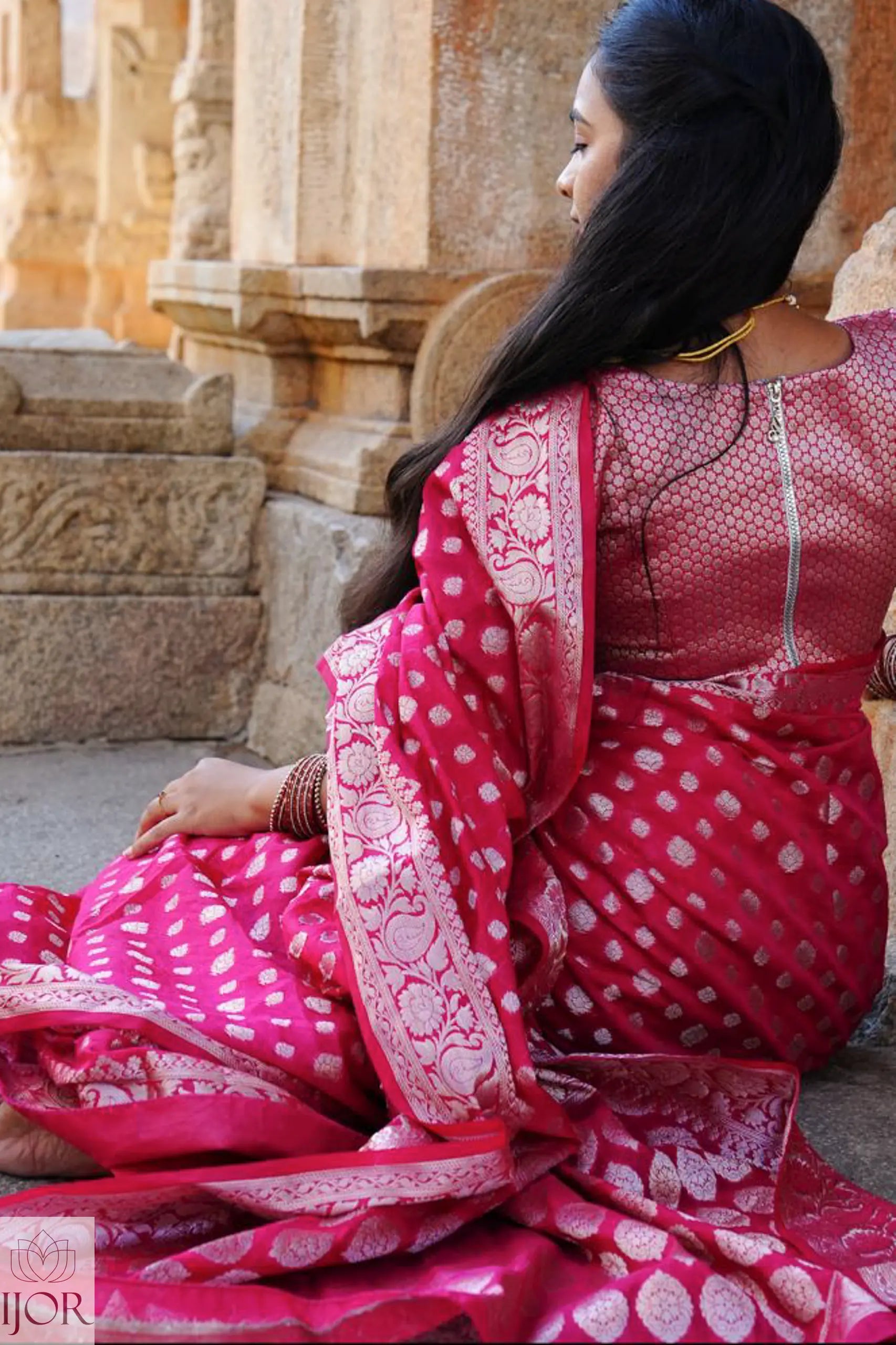 Nandini Yallareddy in Banarasi Soft Georgette Silk Red Saree with silver booti showing casing stiched blouse desin of the saree is sitting on stone steps.