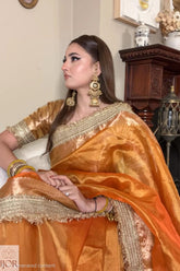 A woman wearing an Banarasi Glass Tissue Silk Saree in Rustic Gold, seated indoors. The saree has a rich orange body with intricate gold zari embroidery along the border and sleeves.