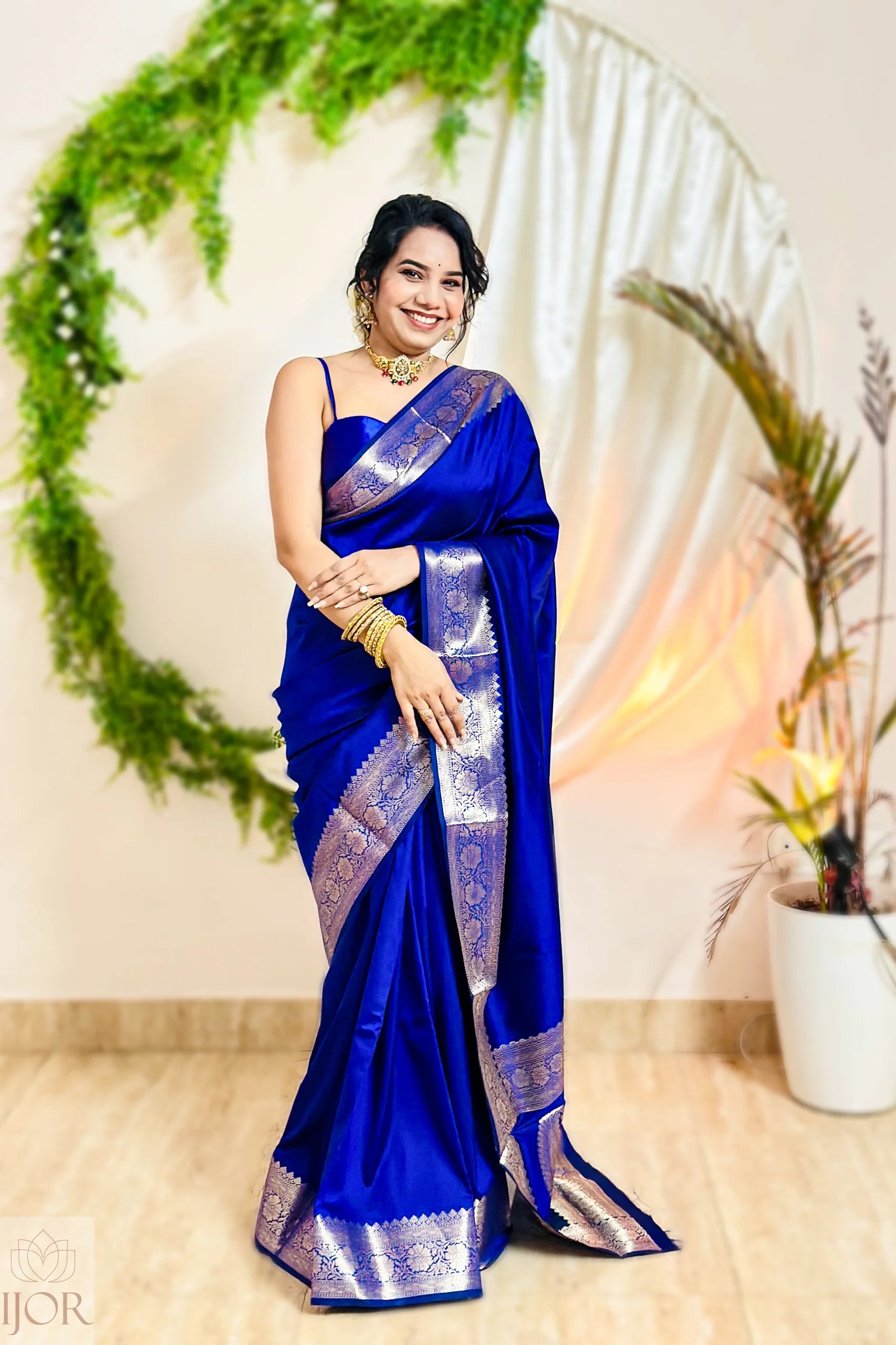 Woman in a blue saree with silver border standing in front of a decorative background.