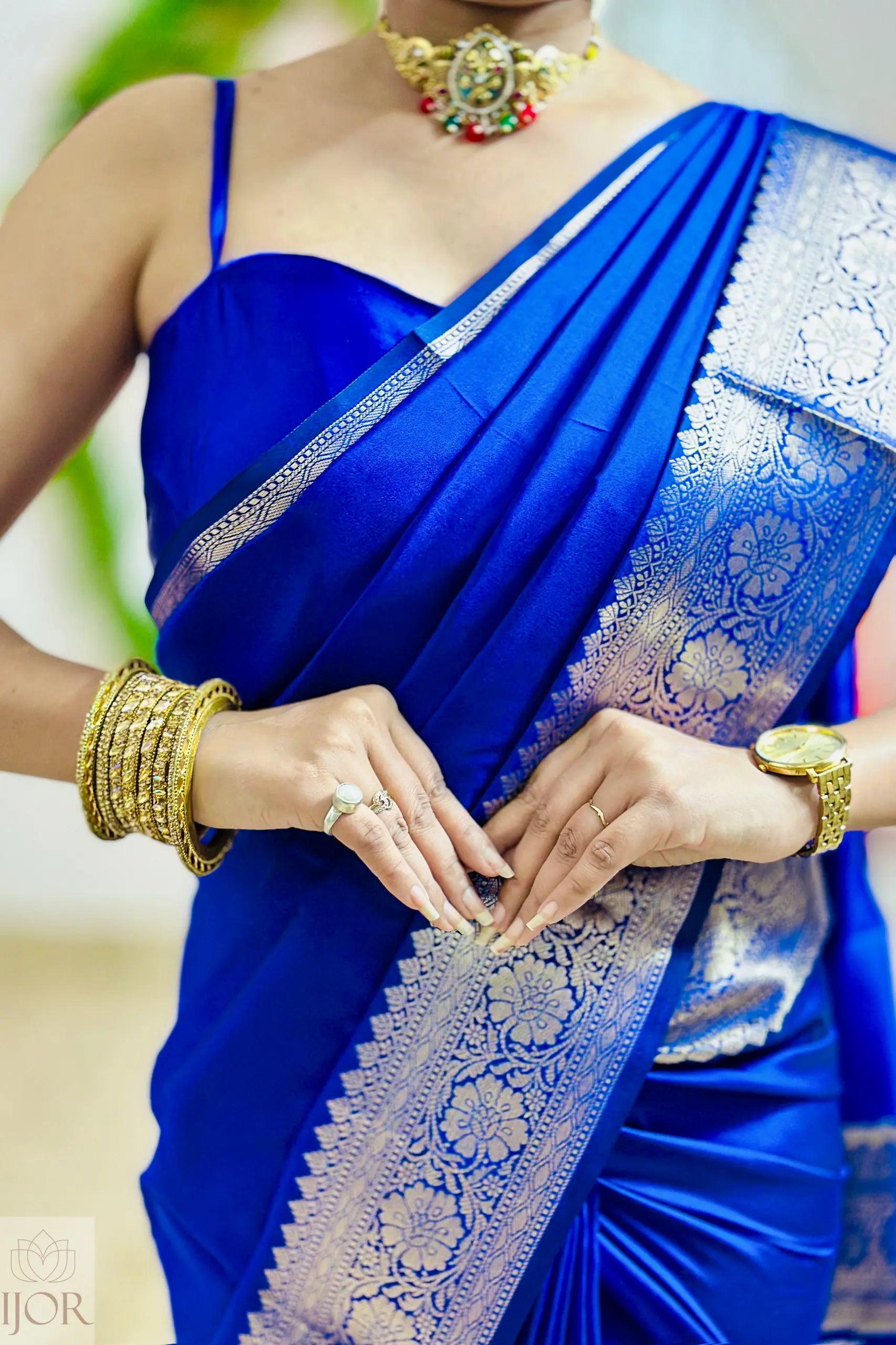 Woman wearing a blue saree with intricate patterns, gold jewelry, and a blurred background.