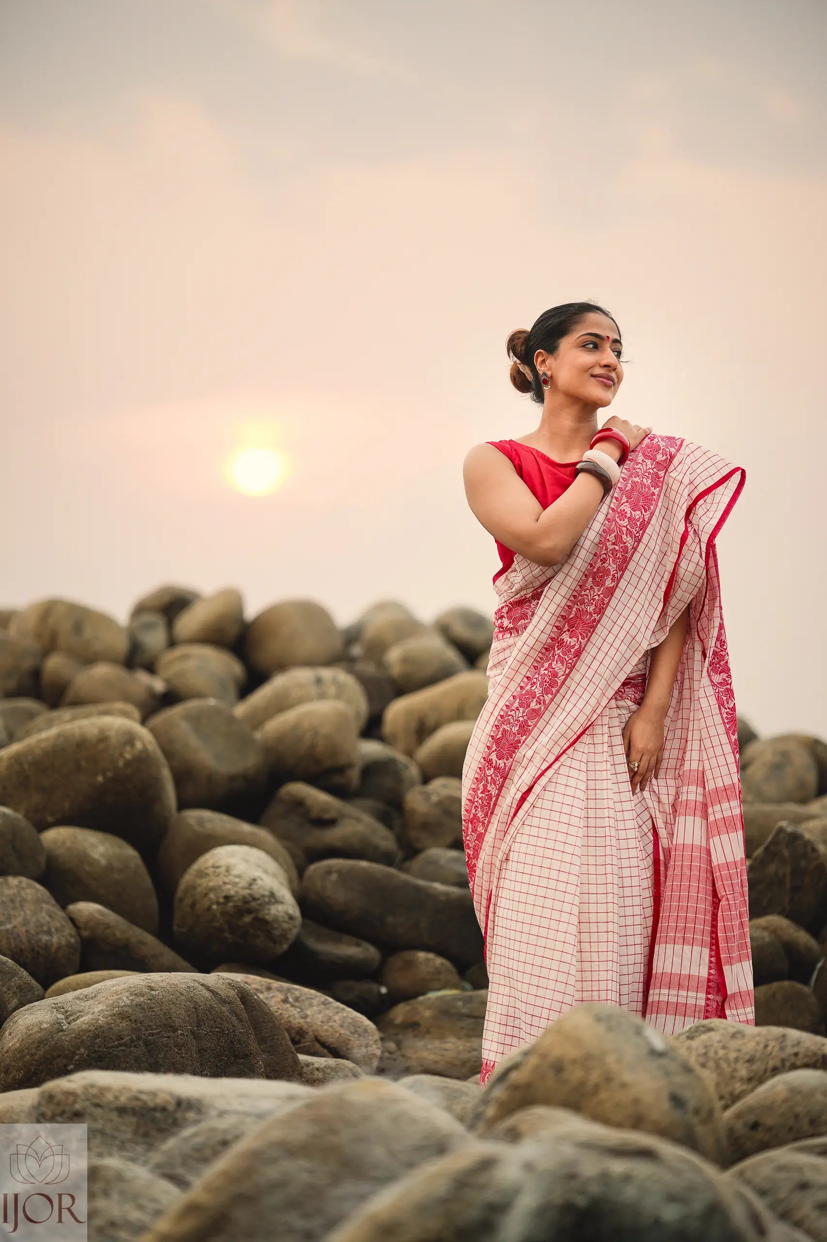 Woman in a Dhonkhali Red and White Pure Cotton Check saree standing among rocks with a sunset in the background