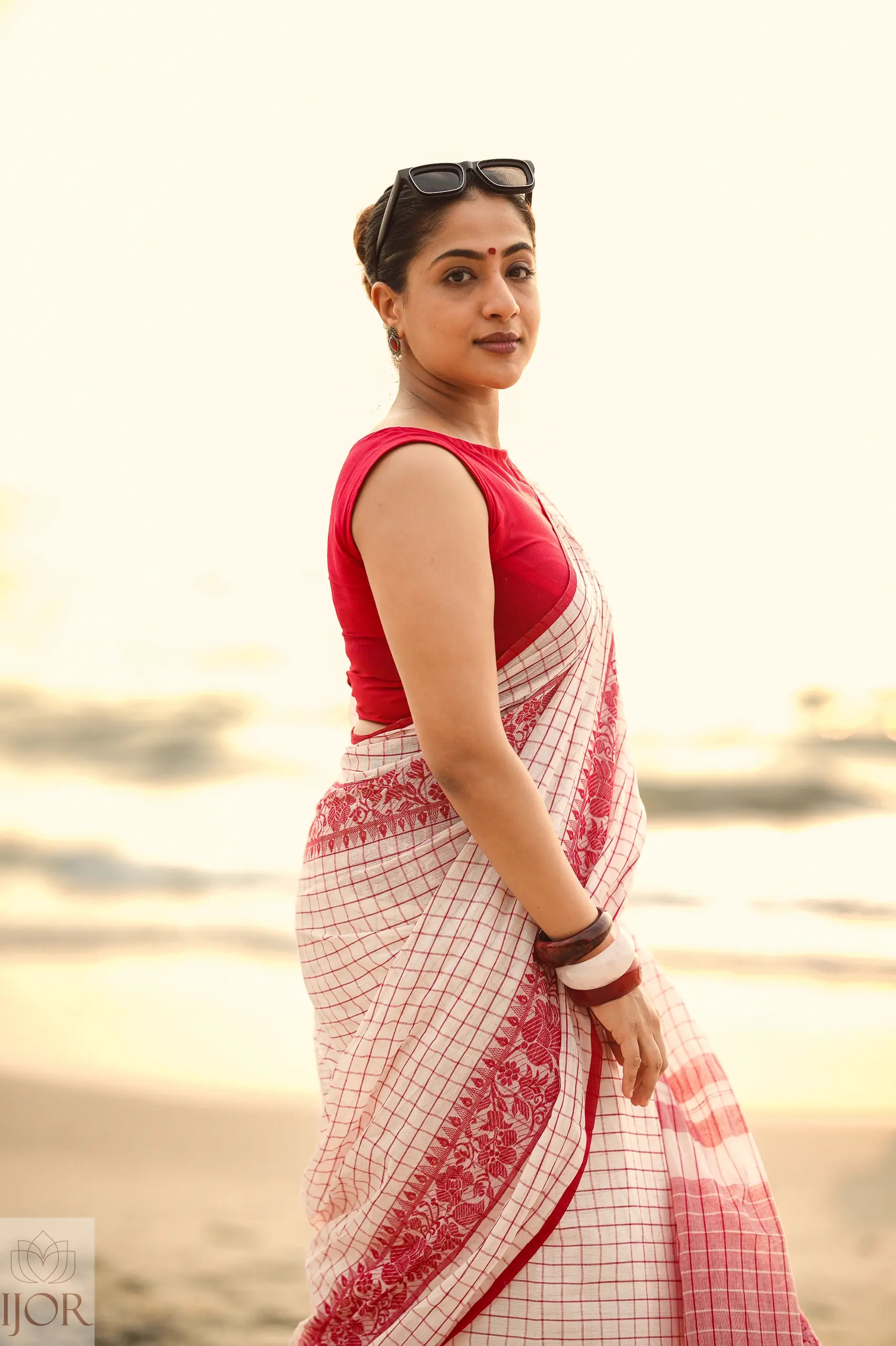 Woman in a Dhonkhali Red and White Pure Cotton Check saree standing by a body of water.