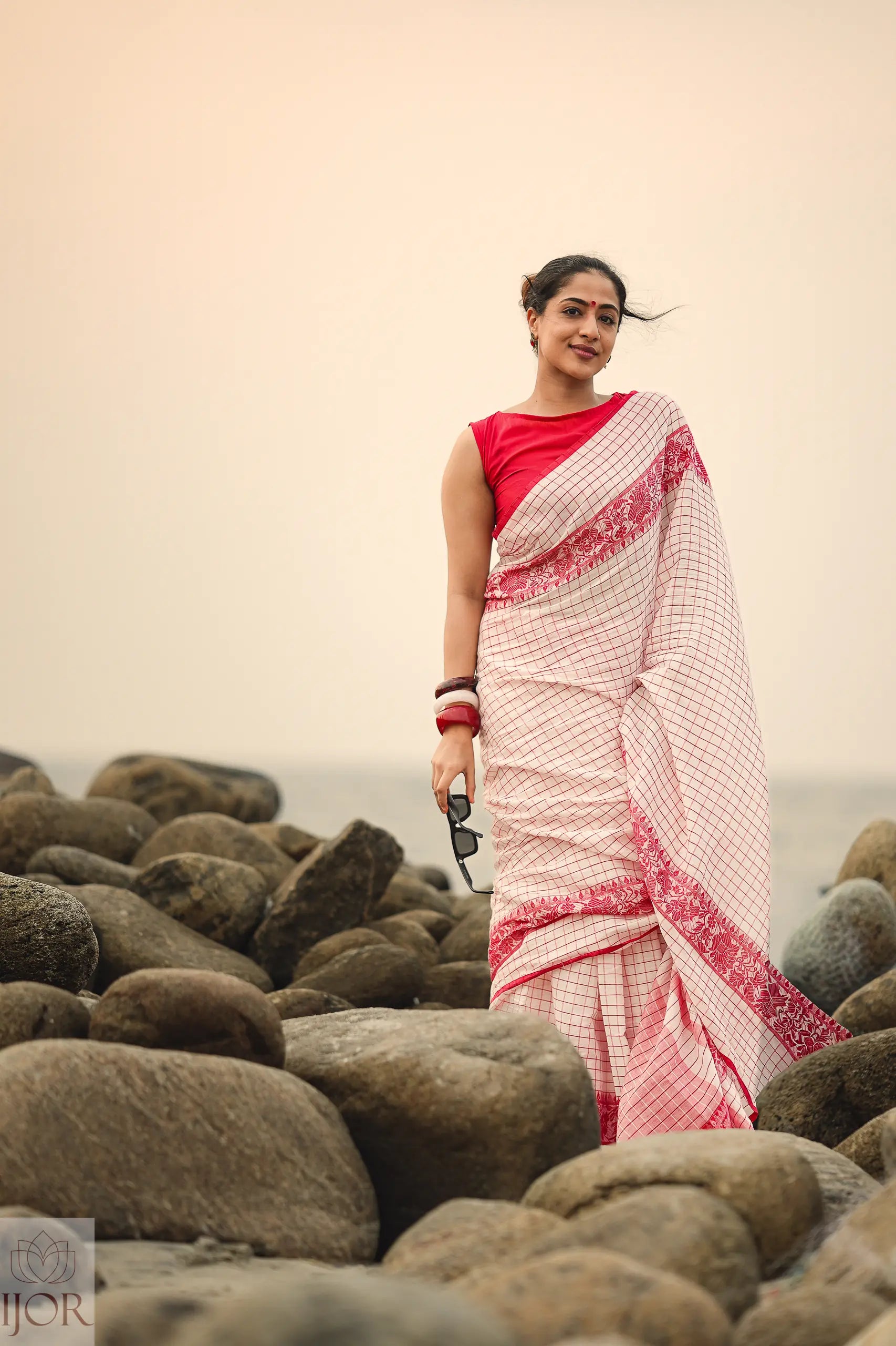 Woman in a Dhonkhali Red and White Pure Cotton Check saree standing on rocks with a blurred background