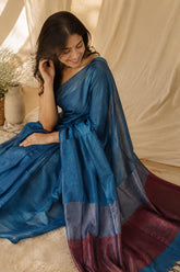 A woman wearing an Blue Pure Khadi Cotton Saree with Zari Anchal with a maroon border, seated in a warm, draped studio setting with beige fabric and a small vase in the background.