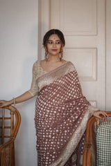 Woman in a Banarasi Soft Georgette Silk Brown Saree standing by a chair against a white wall.