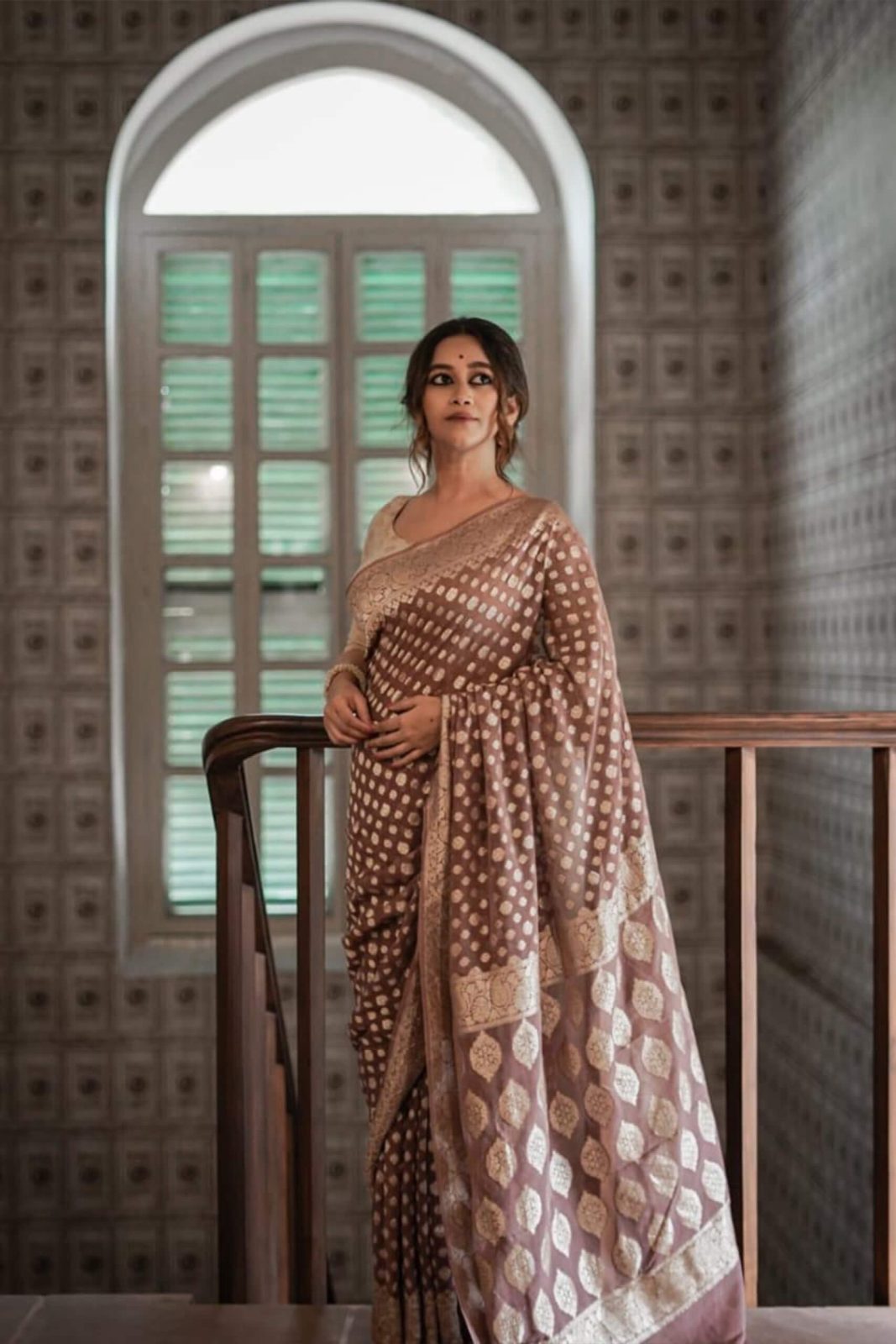Woman in a Banarasi Soft Georgette Silk Brown Saree standing in a room with a decorative wall and window.