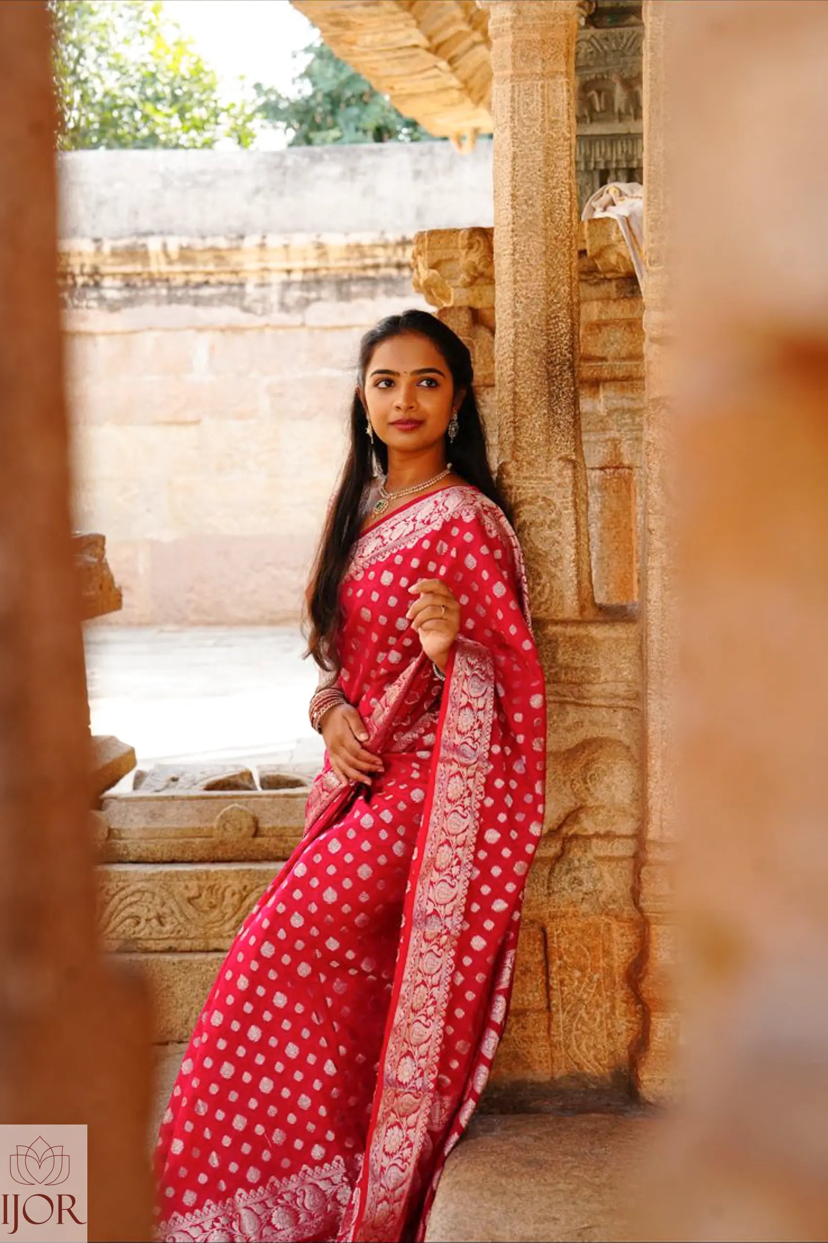 Nandini Yallareddy in Banarasi Soft Georgette Silk Red Saree with silver booti work standing in front of stone architecture.
