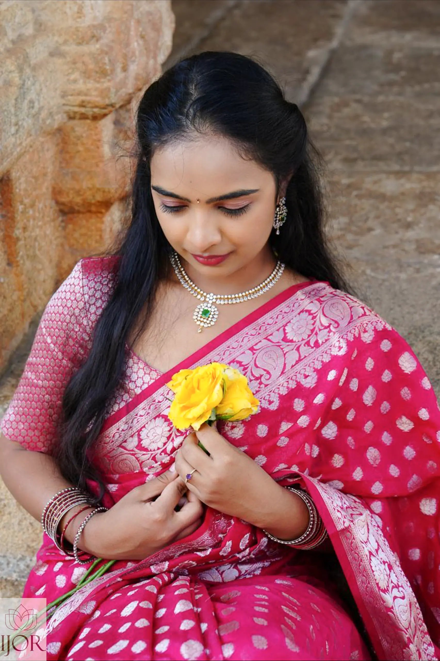 Nandini Yallareddy in Banarasi Soft Georgette Silk Red Saree holding a yellow flower against a stone wall.