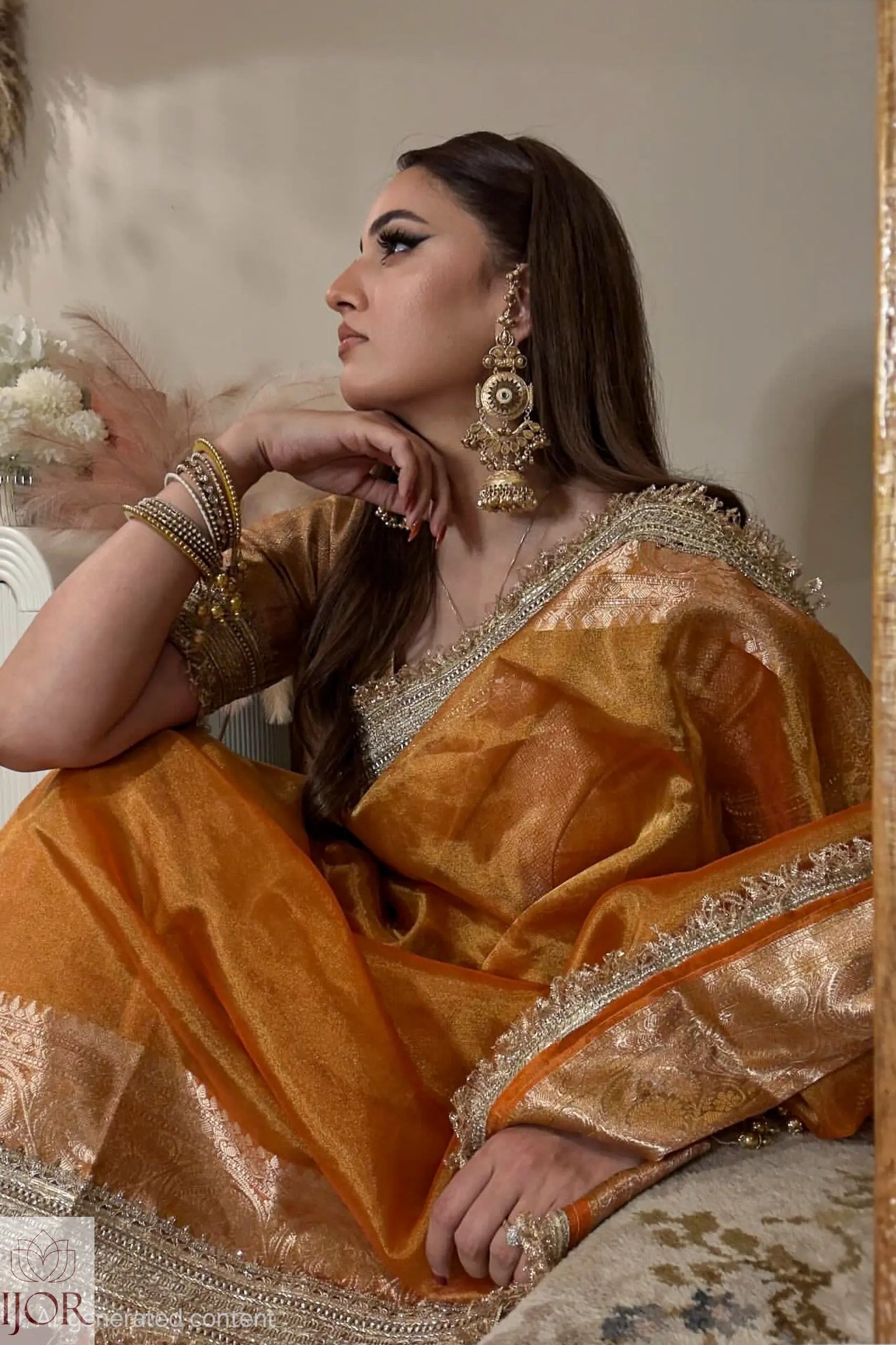 A woman wearing a Banarasi Glass Tissue Silk Saree in Rustic Gold with intricate silver and gold embroidery, styled with traditional Indian jewelry, posing in an indoor setting.
