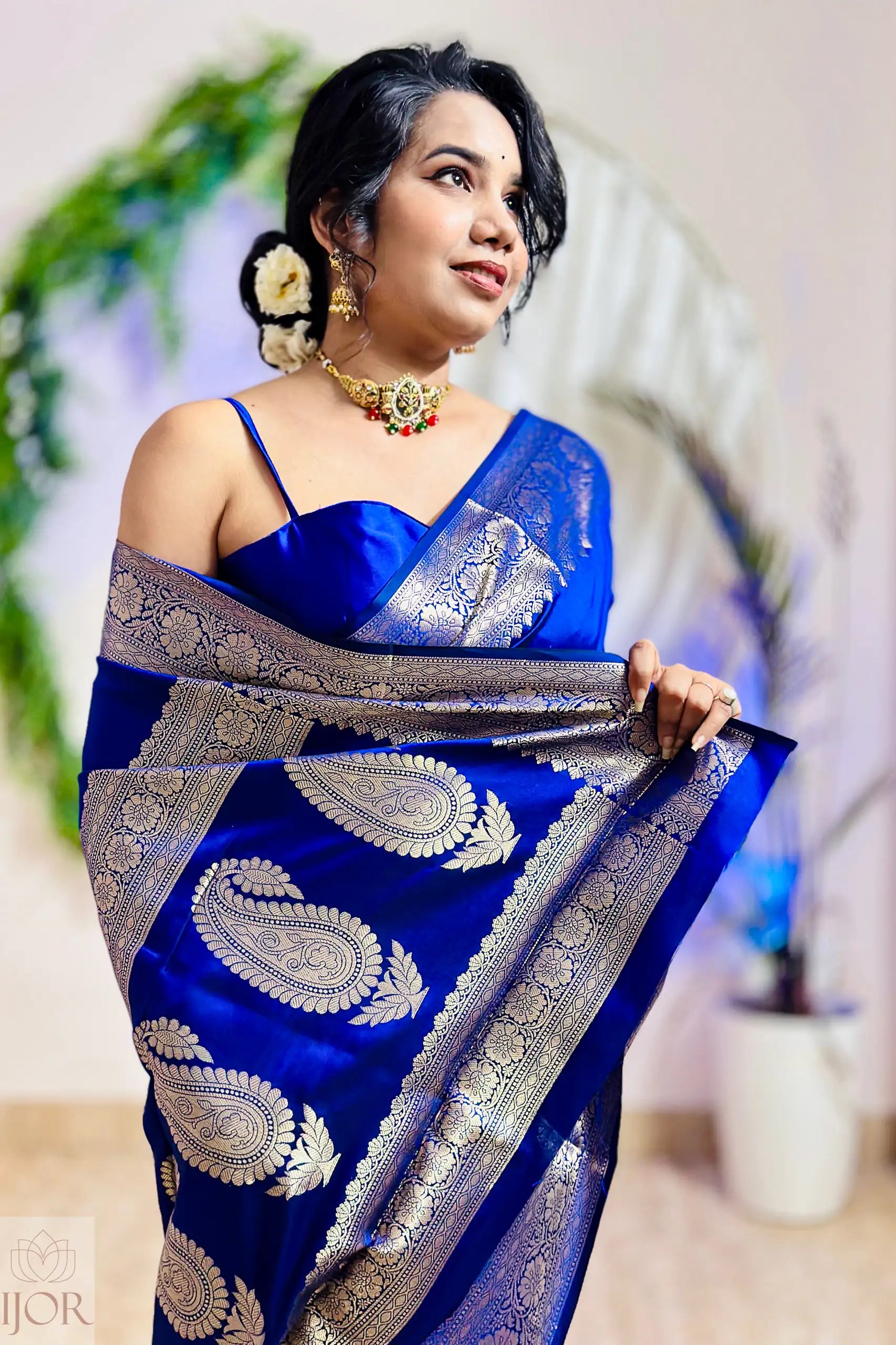 Woman wearing a blue saree with silver patterns, standing indoors with a blurred background.