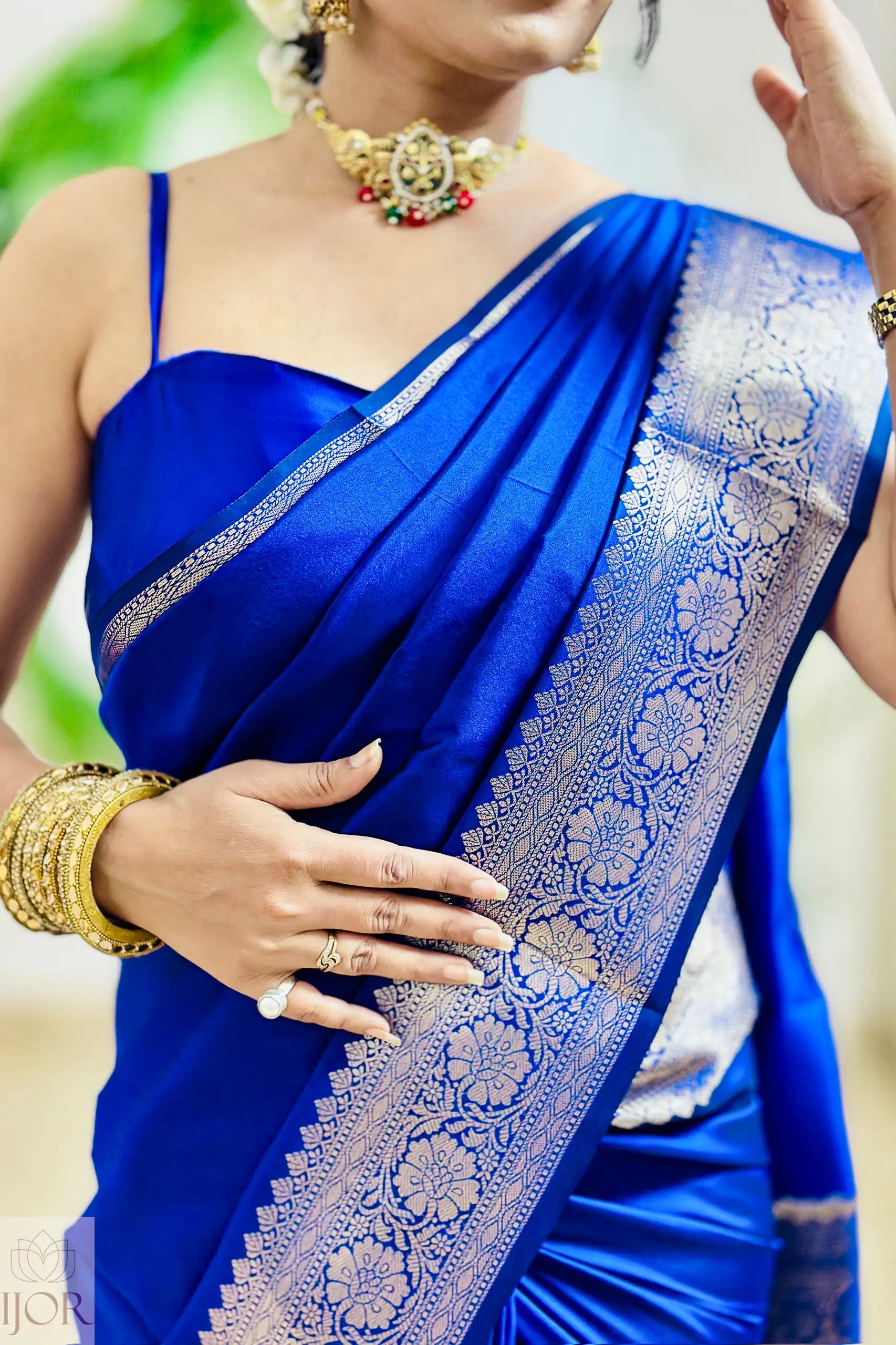 Woman wearing a blue saree with a white border, holding her hand up.