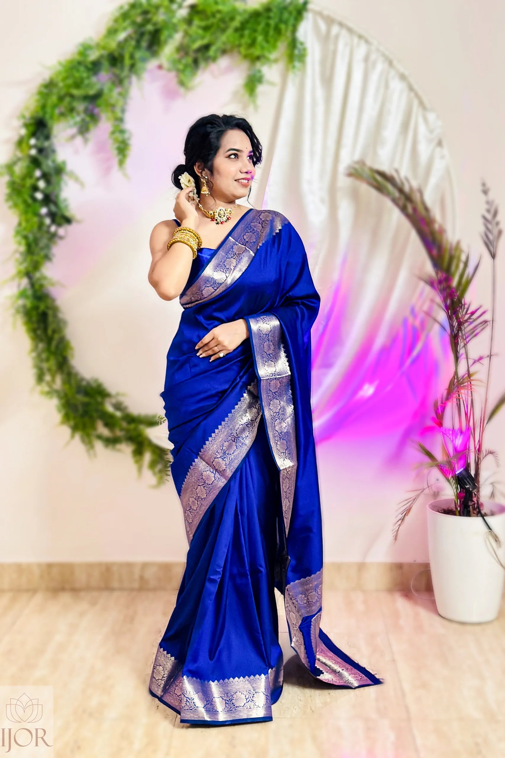 Woman in a blue saree with silver border standing in front of a decorative arch.
