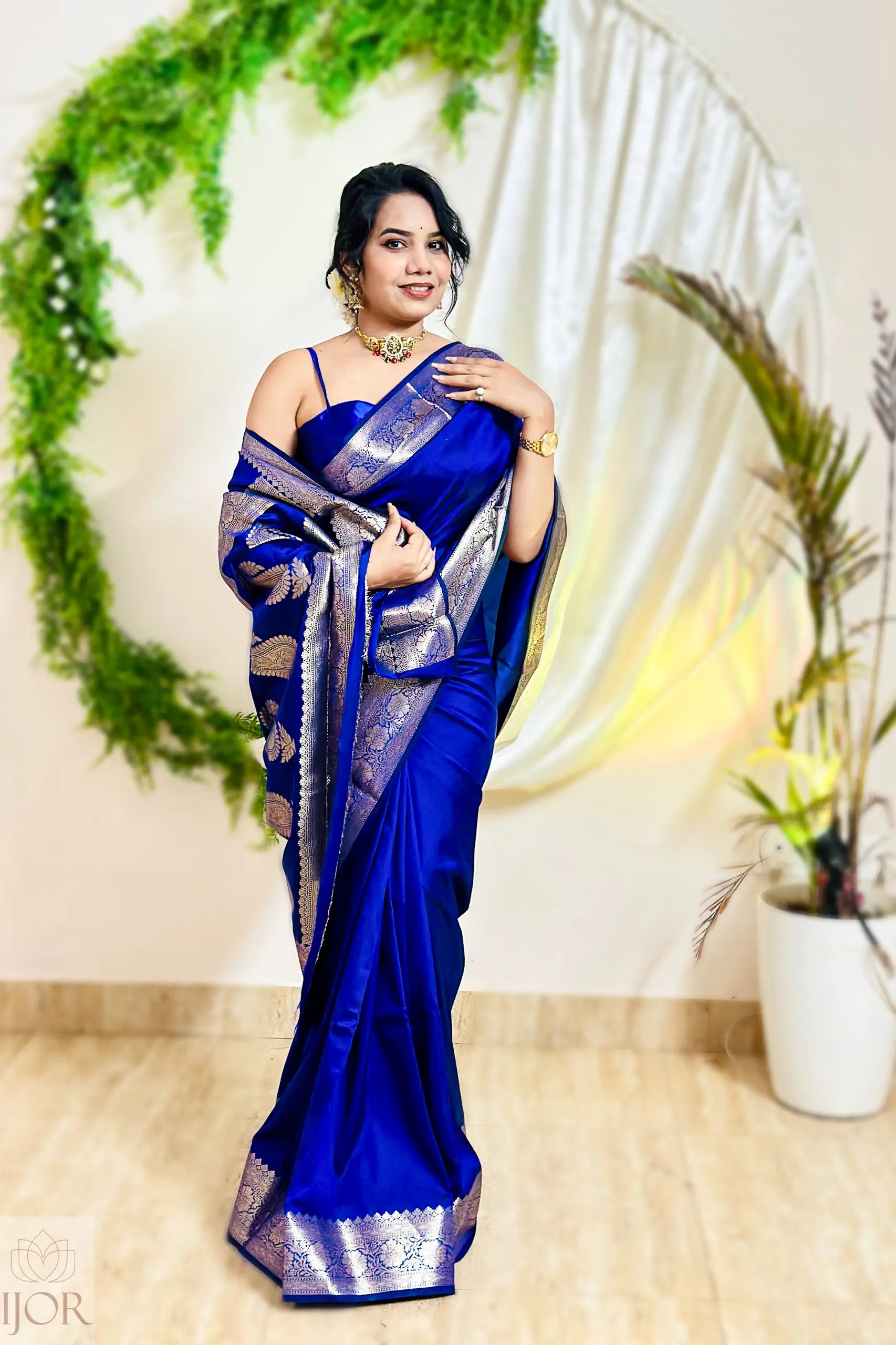 Woman in a blue saree with silver border standing in front of a decorative arch.