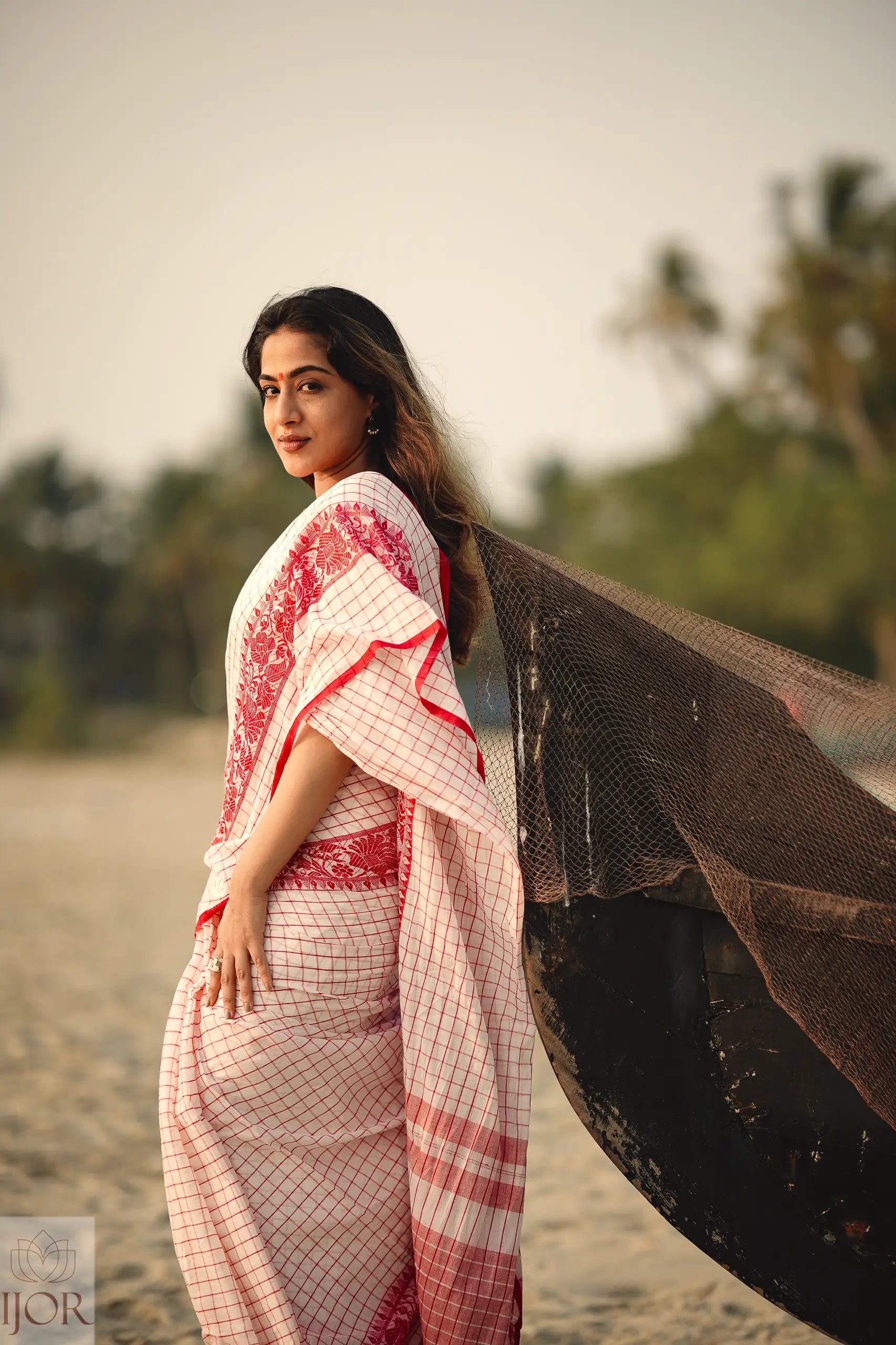 Woman in a Dhonkhali Red and White Pure Cotton Check saree standing on a beach with a blurred background