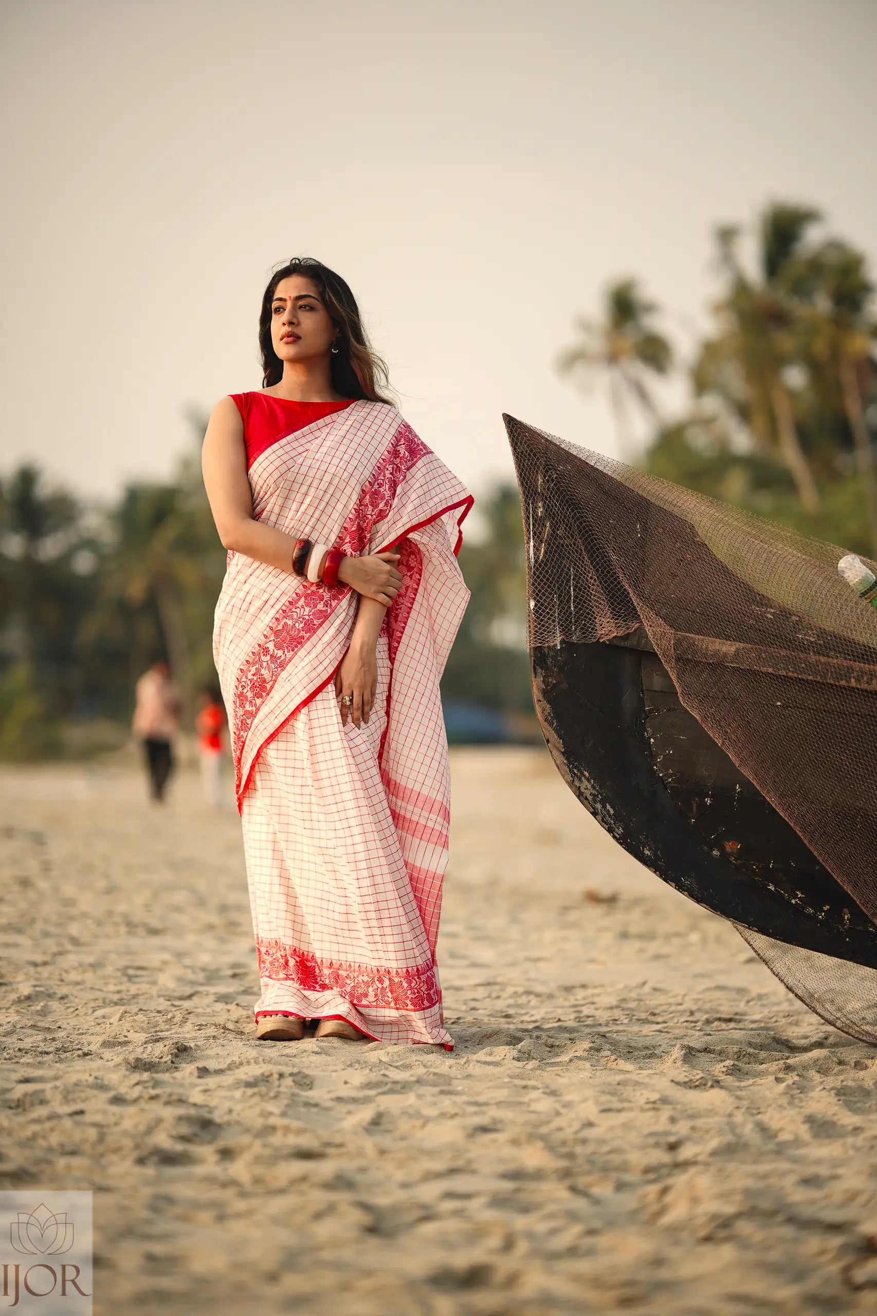 Woman in a Dhonkhali Red and White Pure Cotton Check saree standing on a beach with a traditional boat in the background.