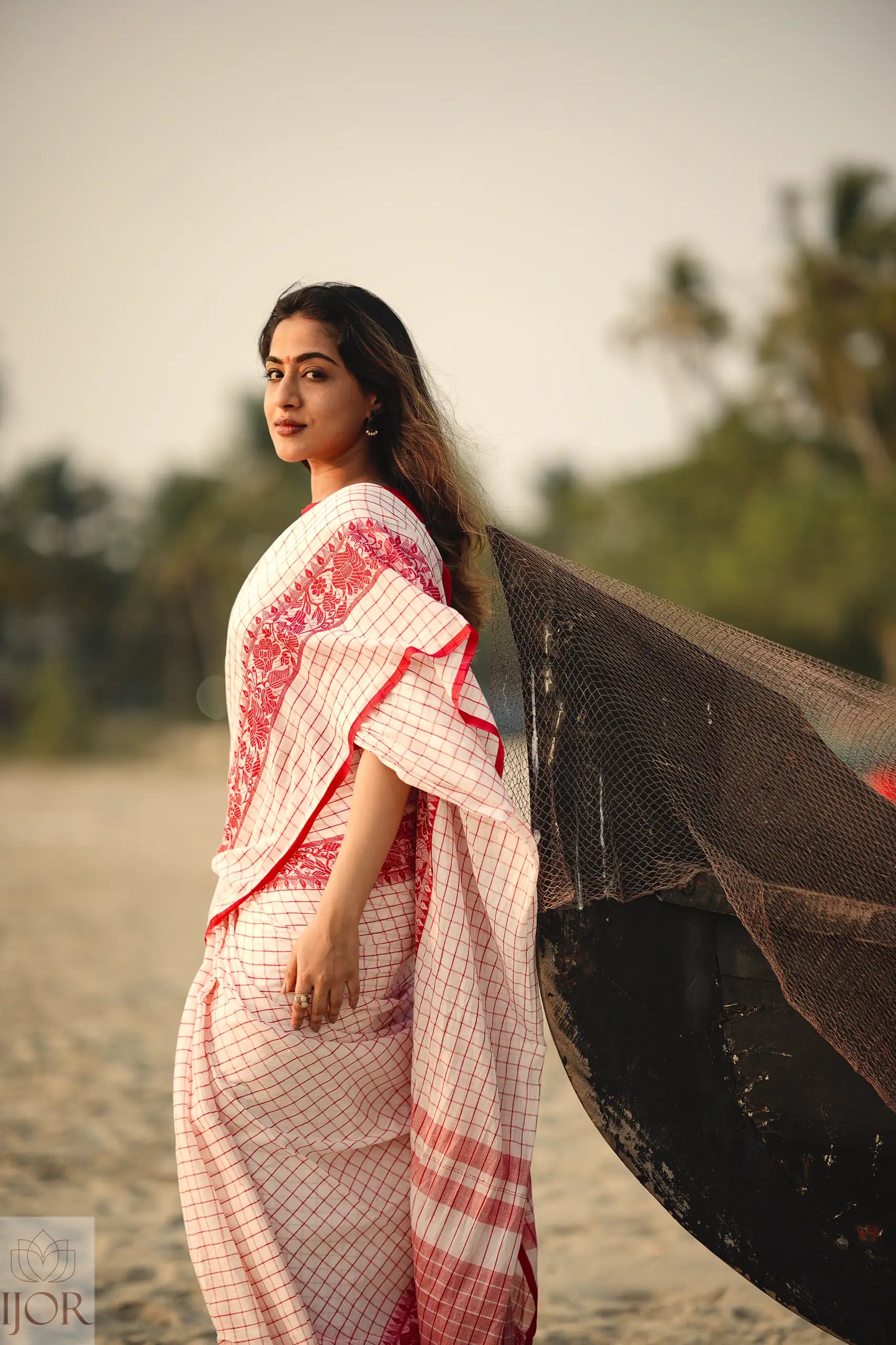 Woman in a Dhonkhali Red and White Pure Cotton Check saree standing next to a fishing net on a beach.