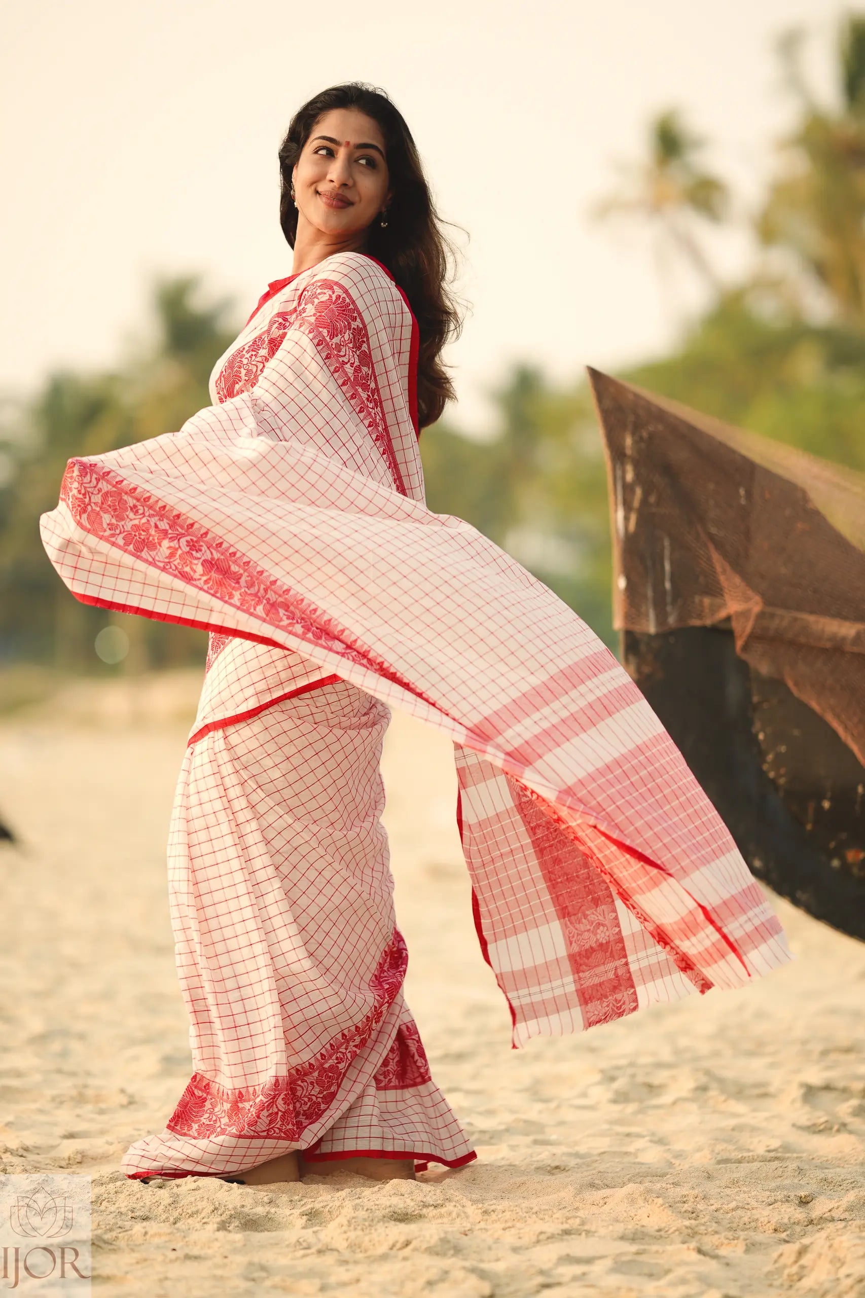 Woman in aDhonkhali Red and White Pure Cotton Check saree standing on a beach with palm trees in the background