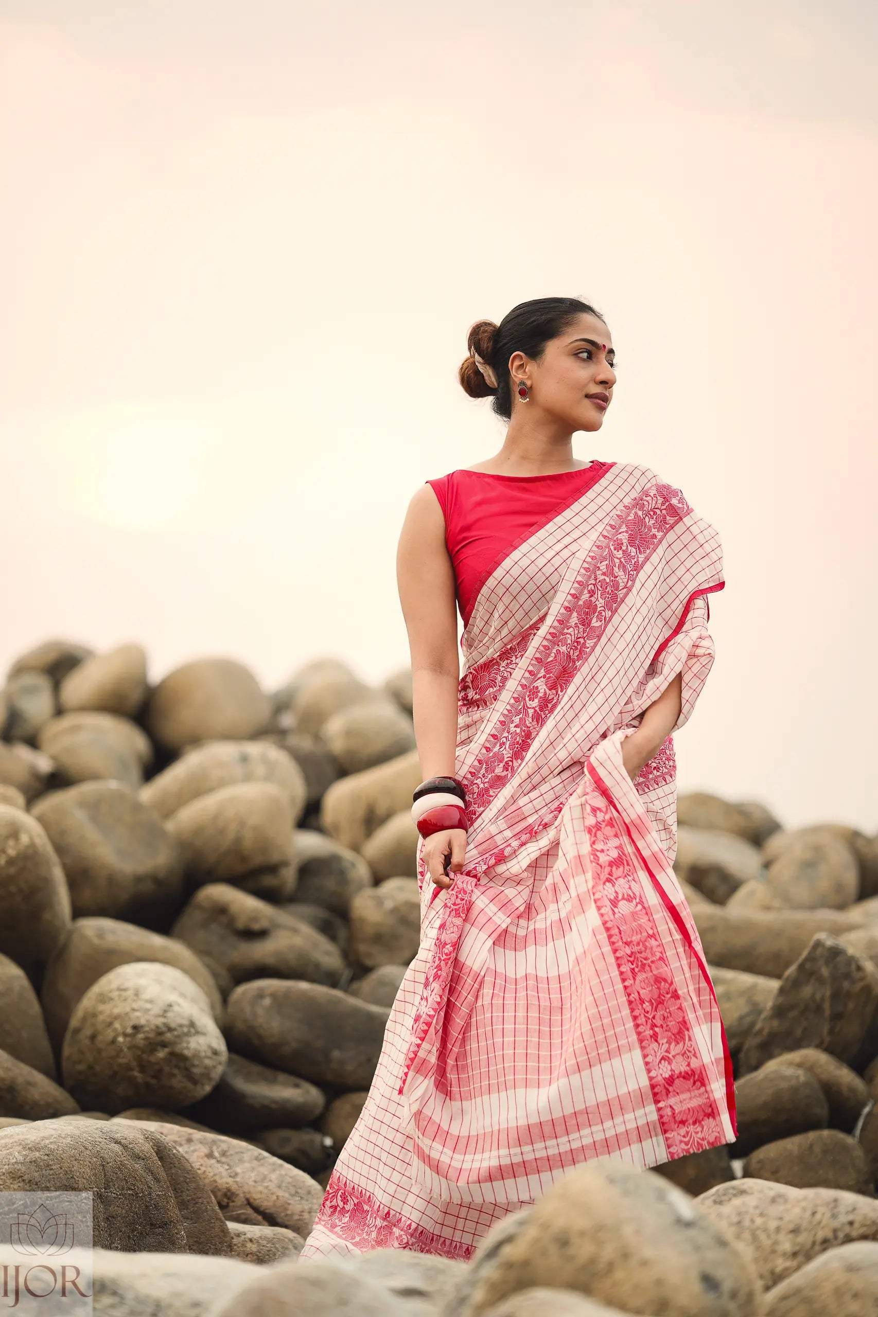 Woman in a Dhonkhali Red and White Pure Cotton Check saree standing among rocks with a blurred background