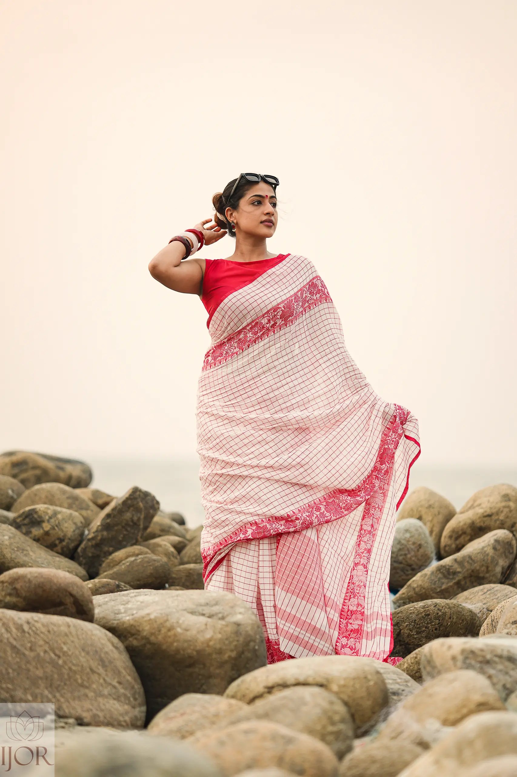 Woman in a Dhonkhali Red and White Pure Cotton Check saree standing among rocks with a plain background