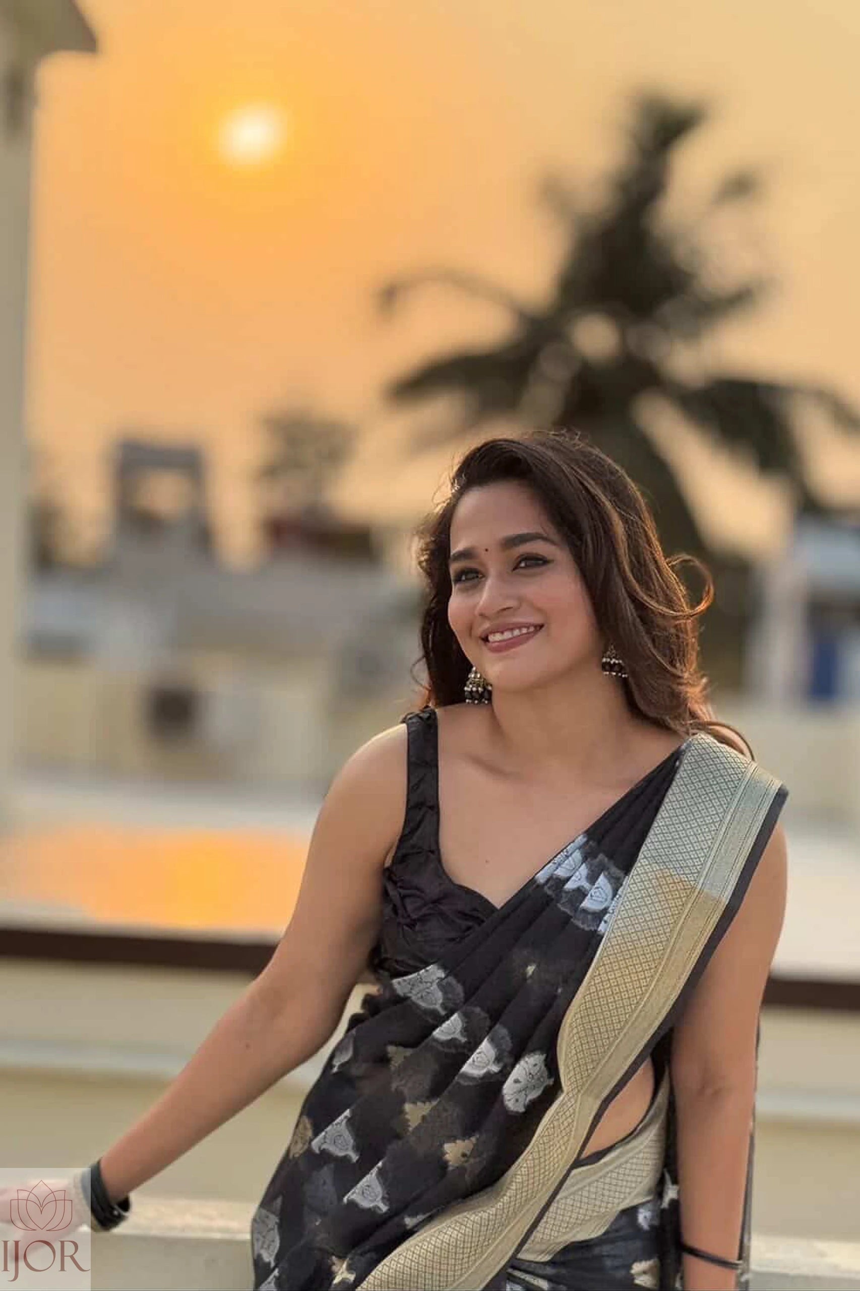 Kavya in a Kora Orgenza Silk Black saree sitting outdoors with palm trees and sunset in the background