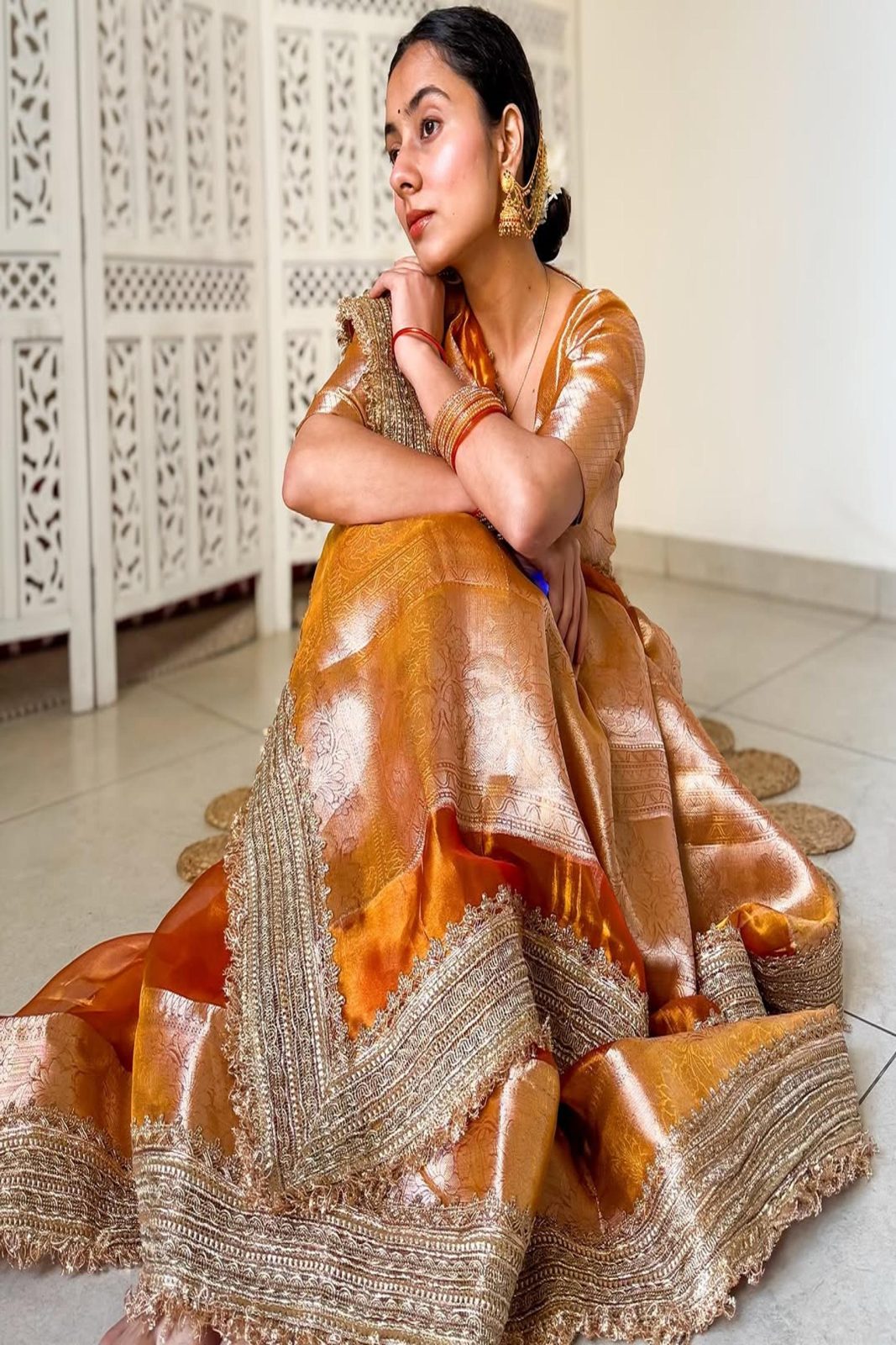 Woman in a traditional orange and gold saree with intricate zari patterns, sitting on a white floor.